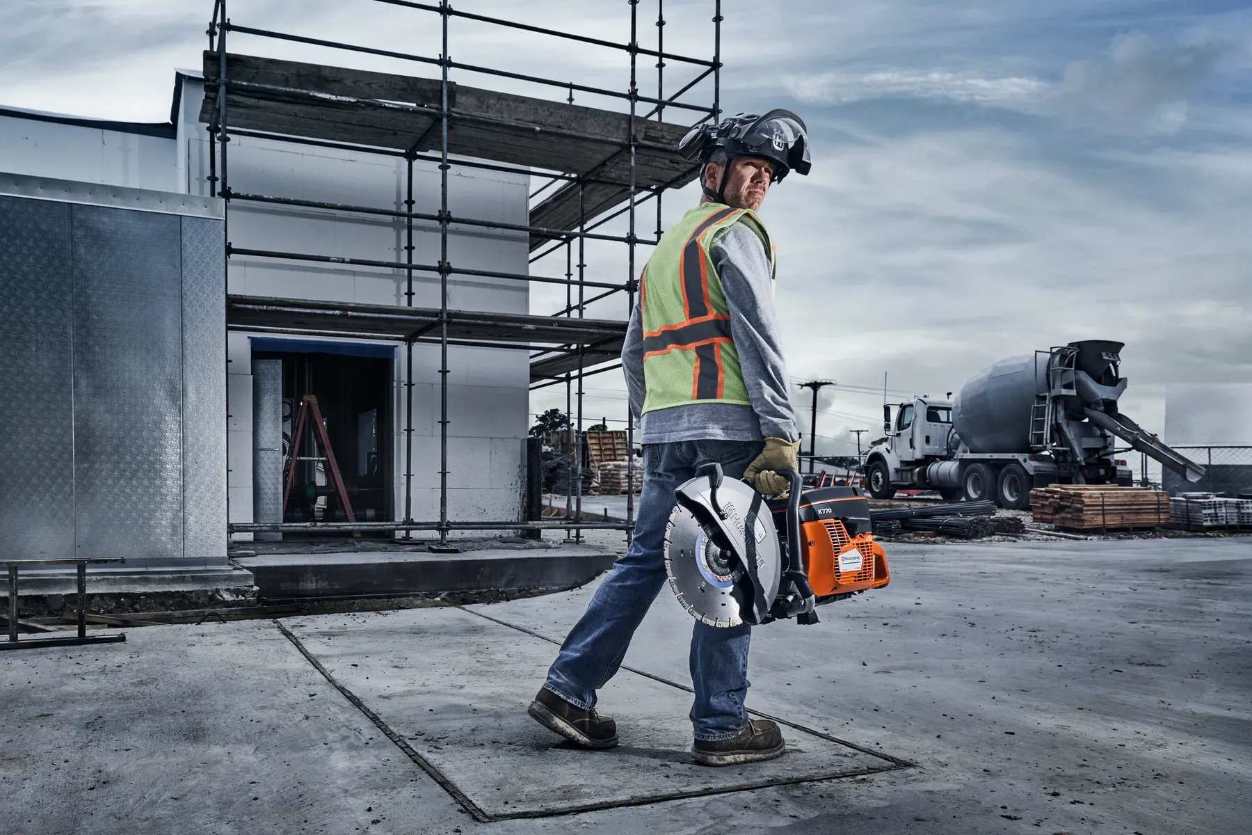 Ouvrier de la construction transportant une découpeuse sur un chantier avec un échafaudage et un camion malaxeur.